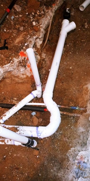 White PVC plumbing pipes laid on red dirt near a wall, with one joint bent into a Y shape.