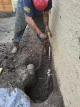 Worker digging a trench beside a brick wall, using a shovel in muddy ground.