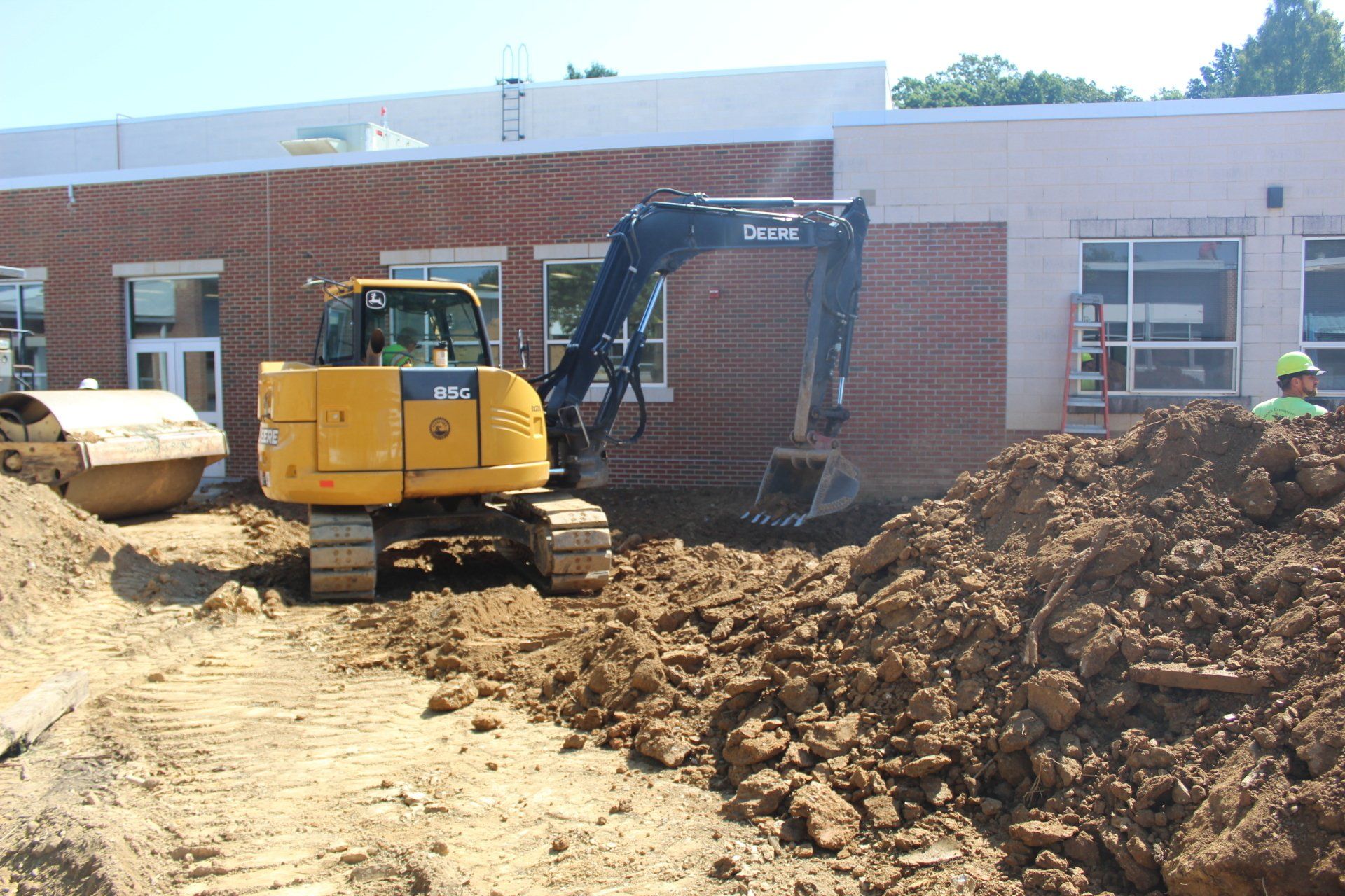 A yellow deere excavator is moving dirt in front of a brick building