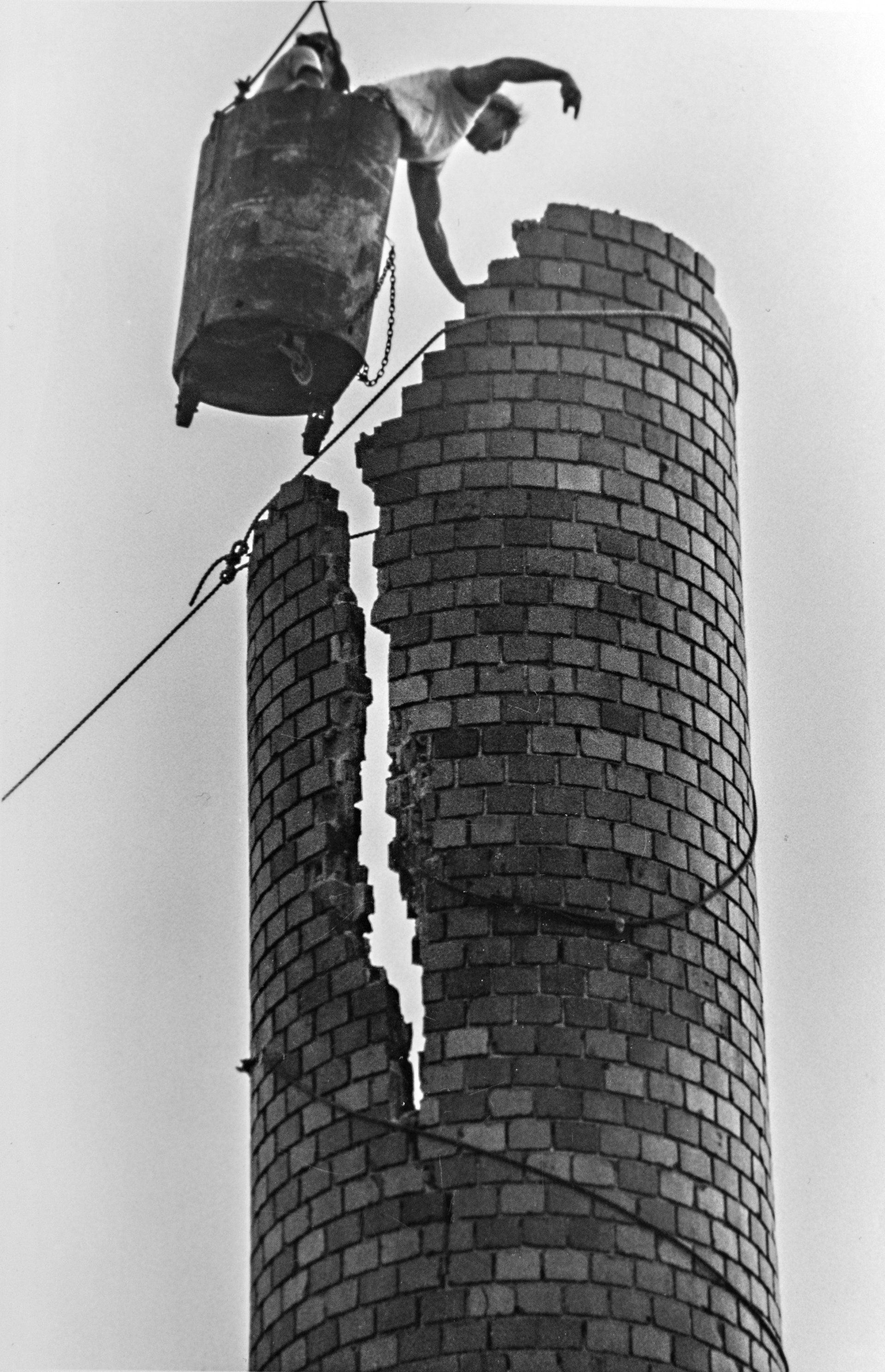 A black and white photo of a man standing on top of a brick chimney.