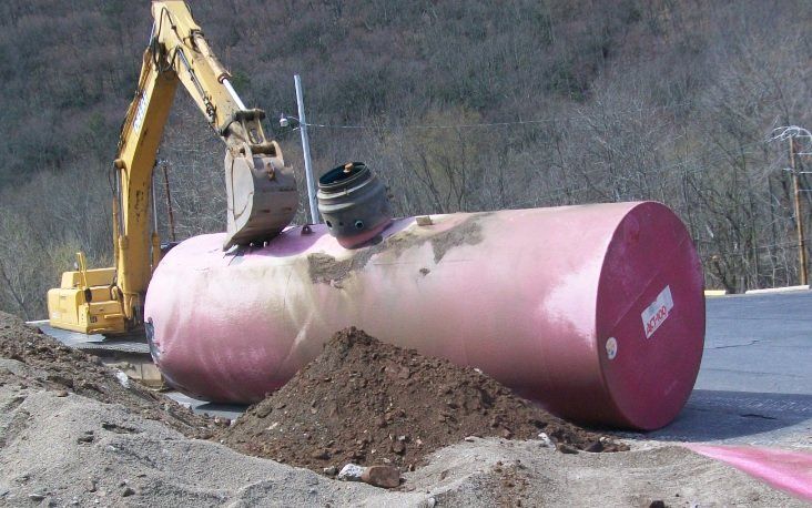 A large red tank is sitting in the dirt next to a yellow excavator.