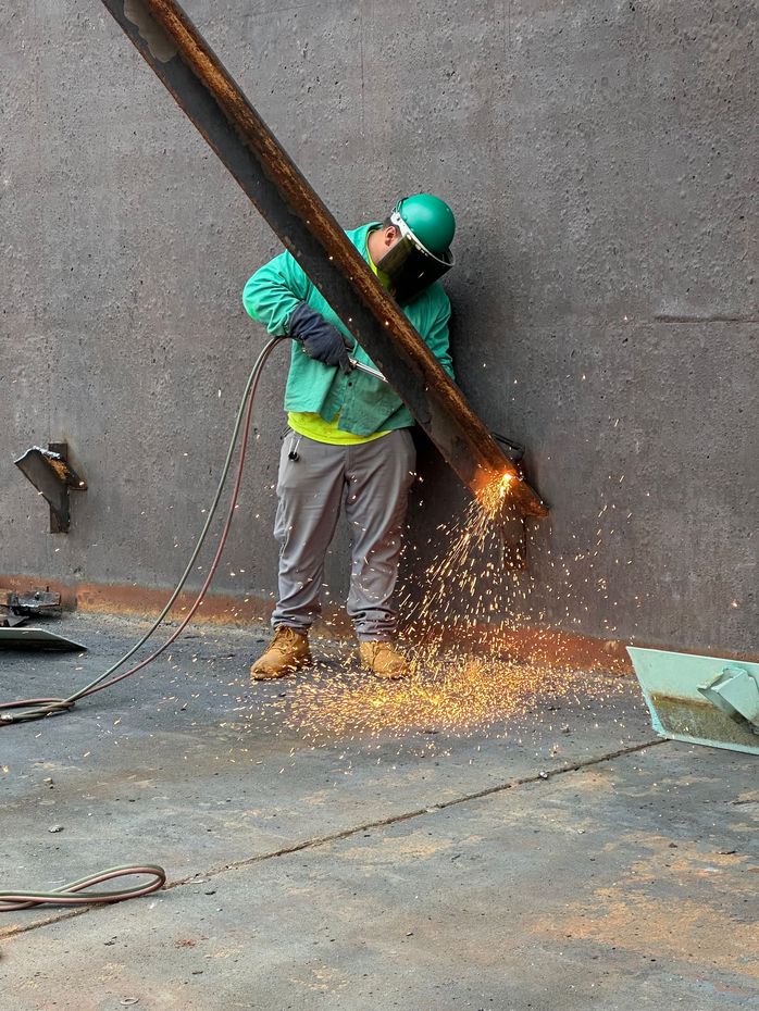 A worker wearing protective gear cuts metal with a torch, generating sparks.