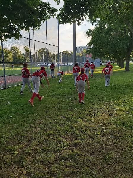 A group of youth baseball players in red jerseys and white pants walking across a grassy field near a chain-link fence.