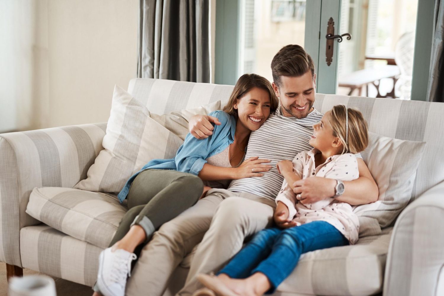 Family of three, a smiling woman, man, and child, cuddle on a sofa in a living room.