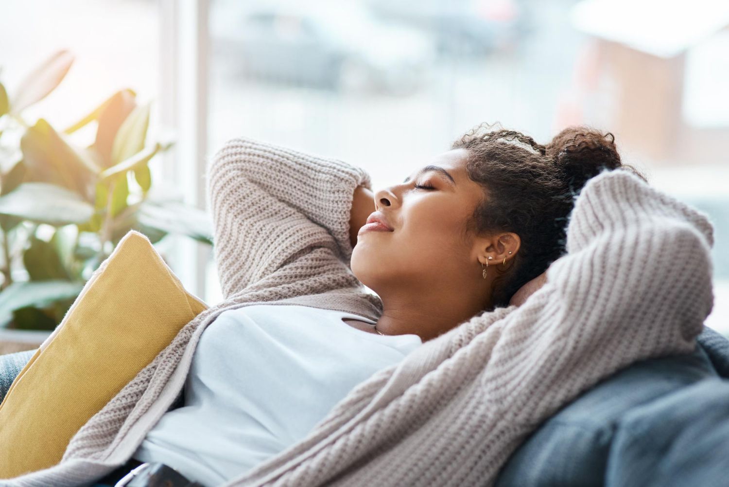 Woman relaxing on a couch, hands behind head, eyes closed. Sunlit room, window in the background.