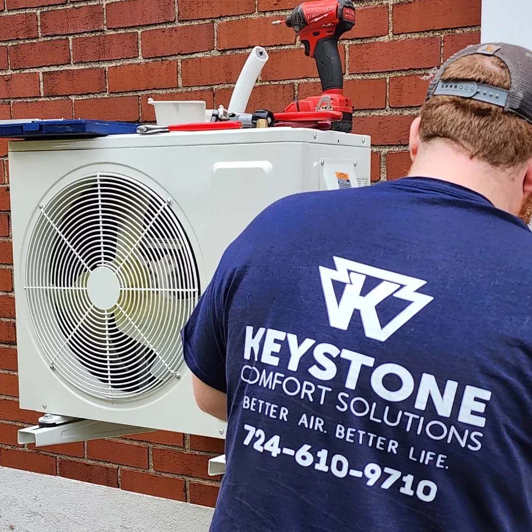 HVAC technician installing an air conditioning unit on a brick wall.  The technician wears a blue shirt with the Keystone Comfort Solutions logo.