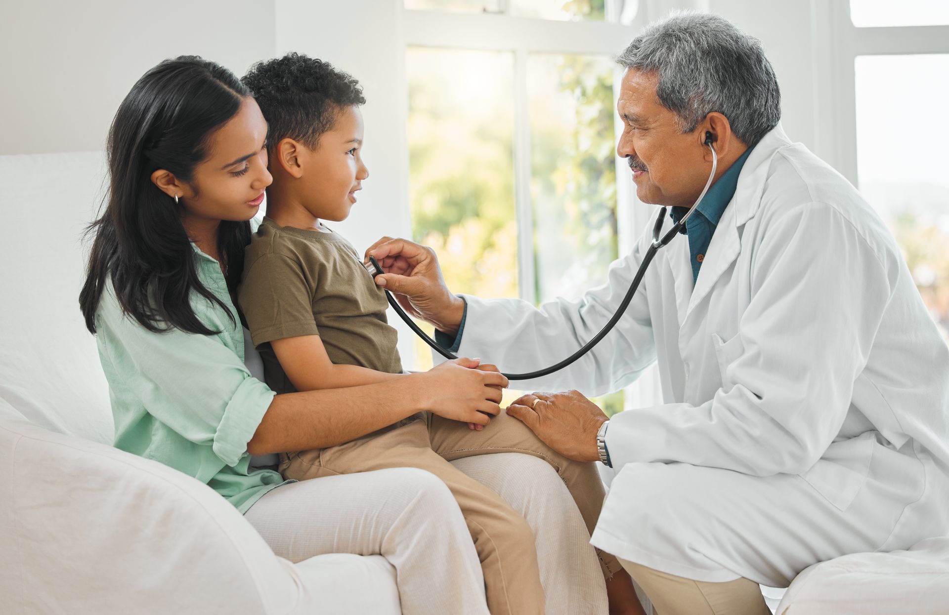 Woman holding child on lap with doctor checking child with stethoscope.