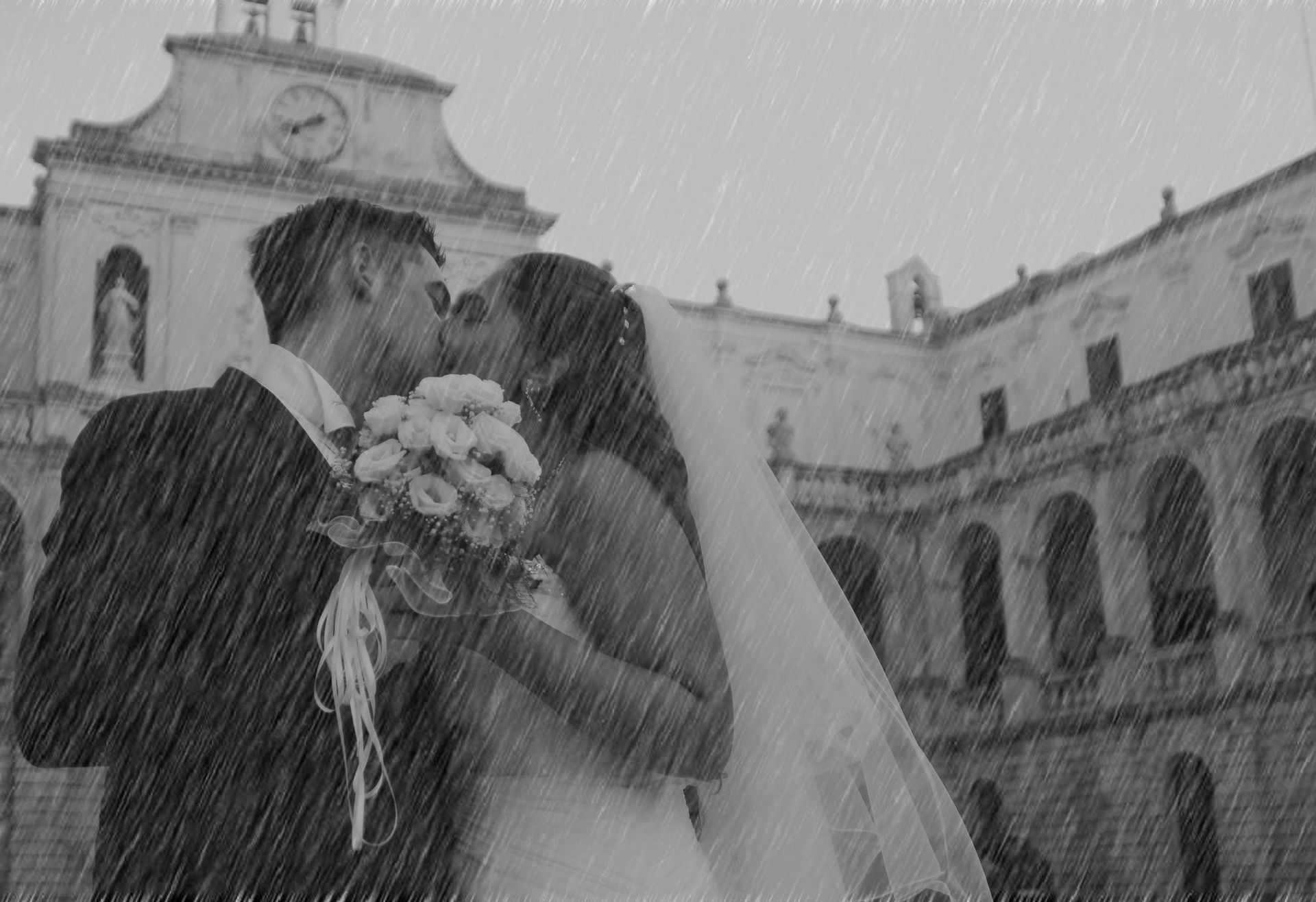 Una sposa e uno sposo che si baciano sotto la pioggia davanti a un edificio.