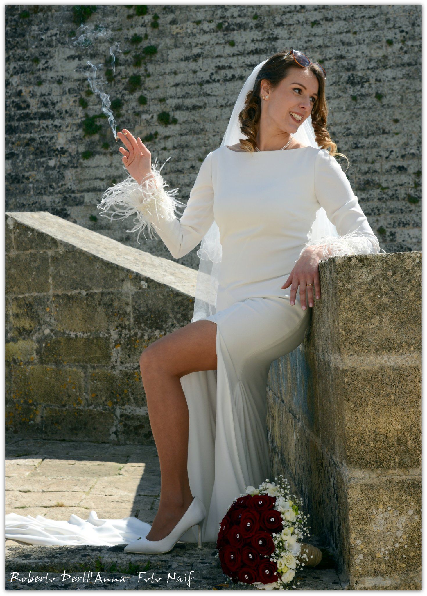 Una donna in abito da sposa è seduta su un muro di pietra e tiene in mano un mazzo di fiori.