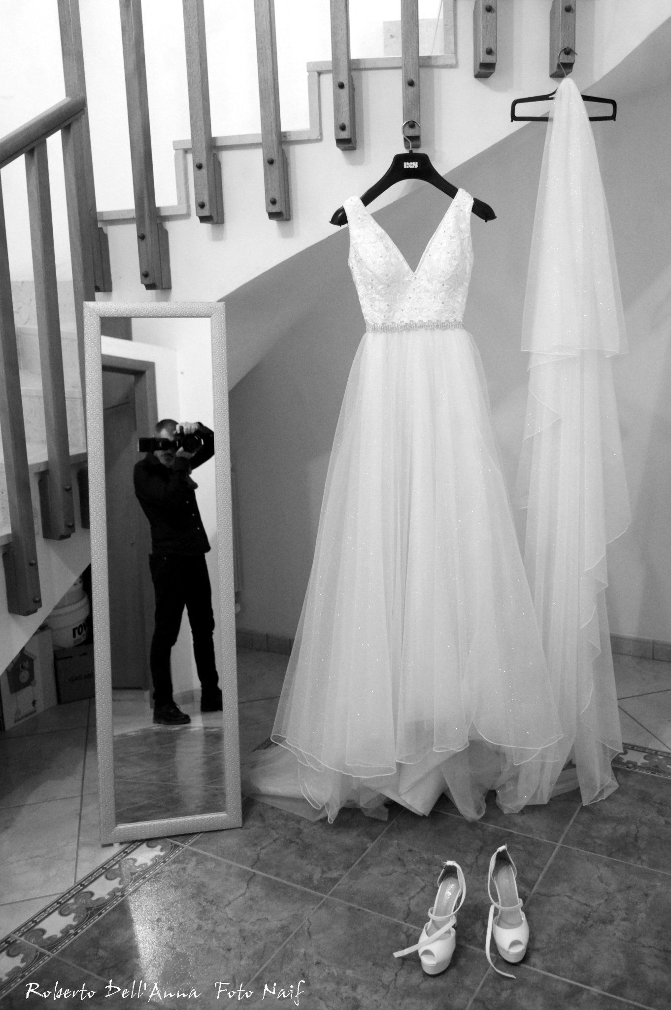 Una foto in bianco e nero di un abito da sposa appeso a una gruccia accanto a uno specchio.