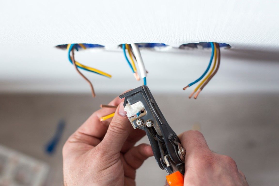 Hands stripping electrical wires from a ceiling junction box with a wire stripper.