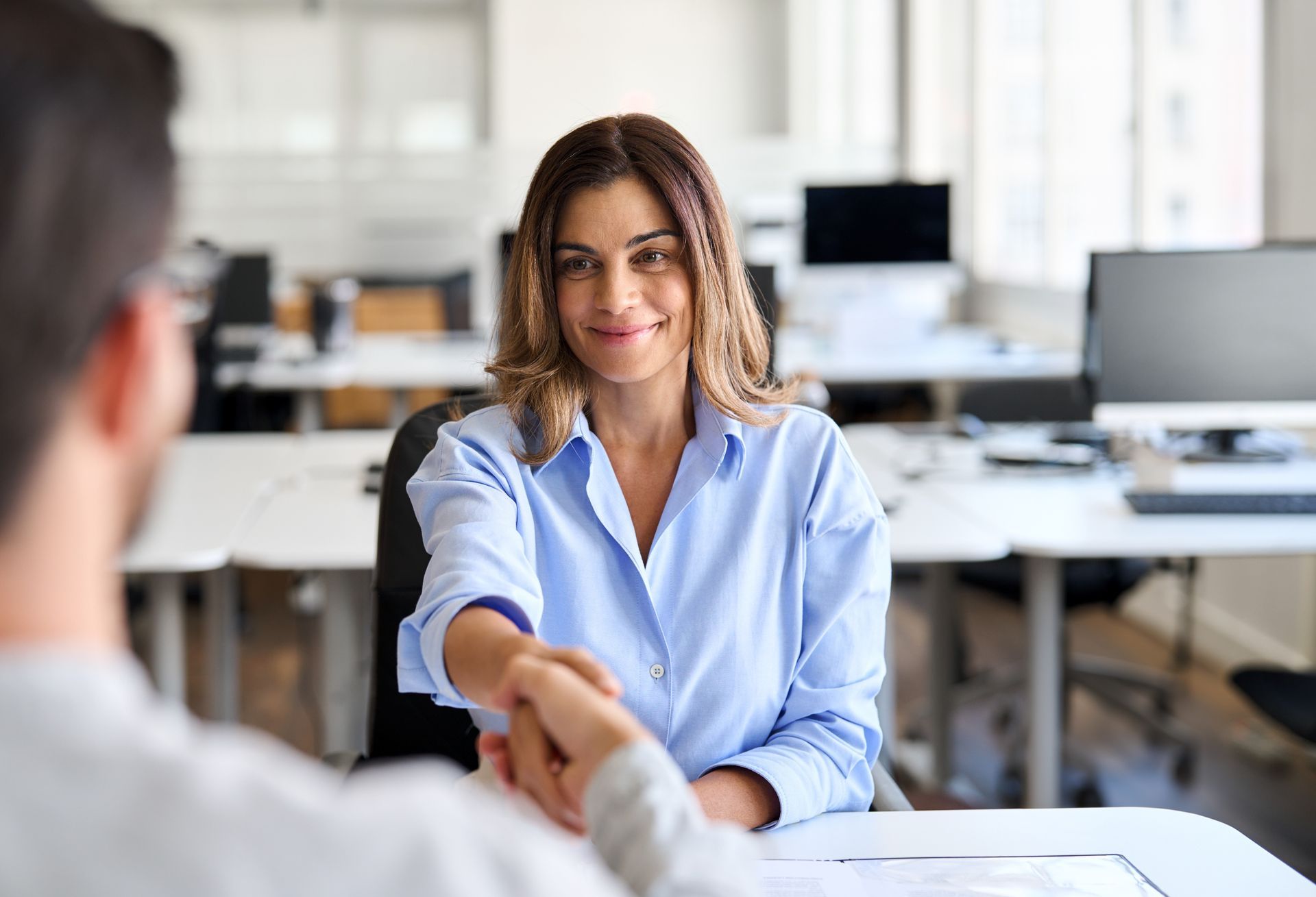 A person in a light blue shirt smiles while shaking hands with someone across a desk in a modern office.
