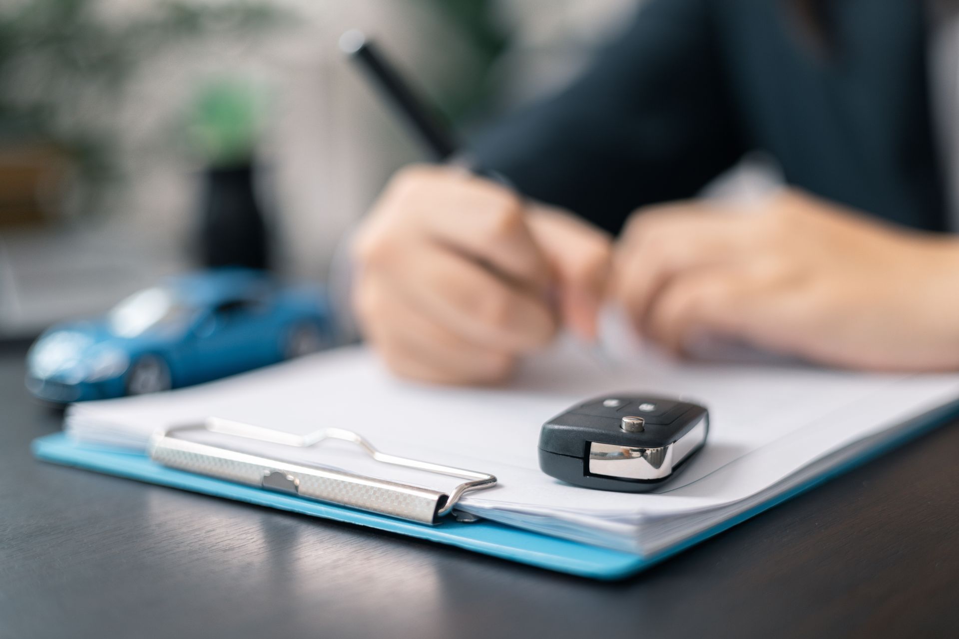 A person signs a document on a clipboard next to a car key fob and a small blue toy car.