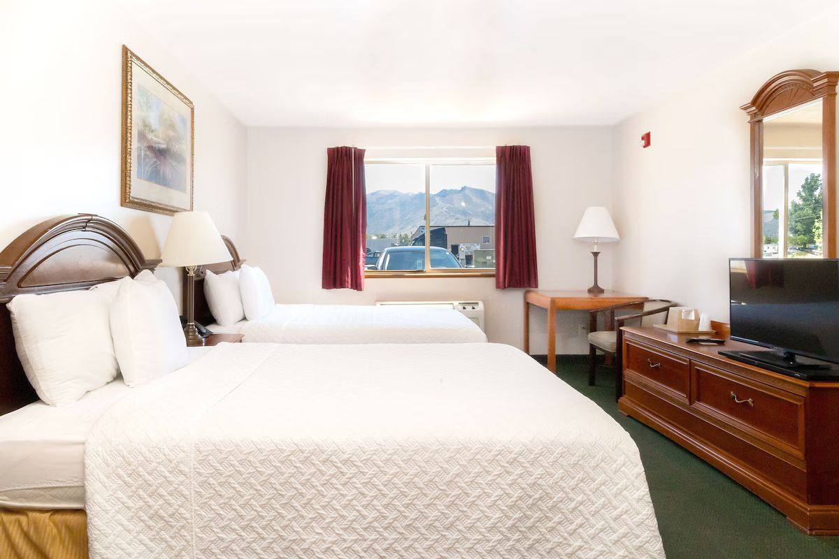 Hotel room with two beds, window with mountain view, dresser with TV, desk.