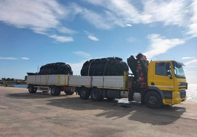Camion giallo con carico coperto da telone parcheggiato in un piazzale aperto sotto un cielo azzurro.