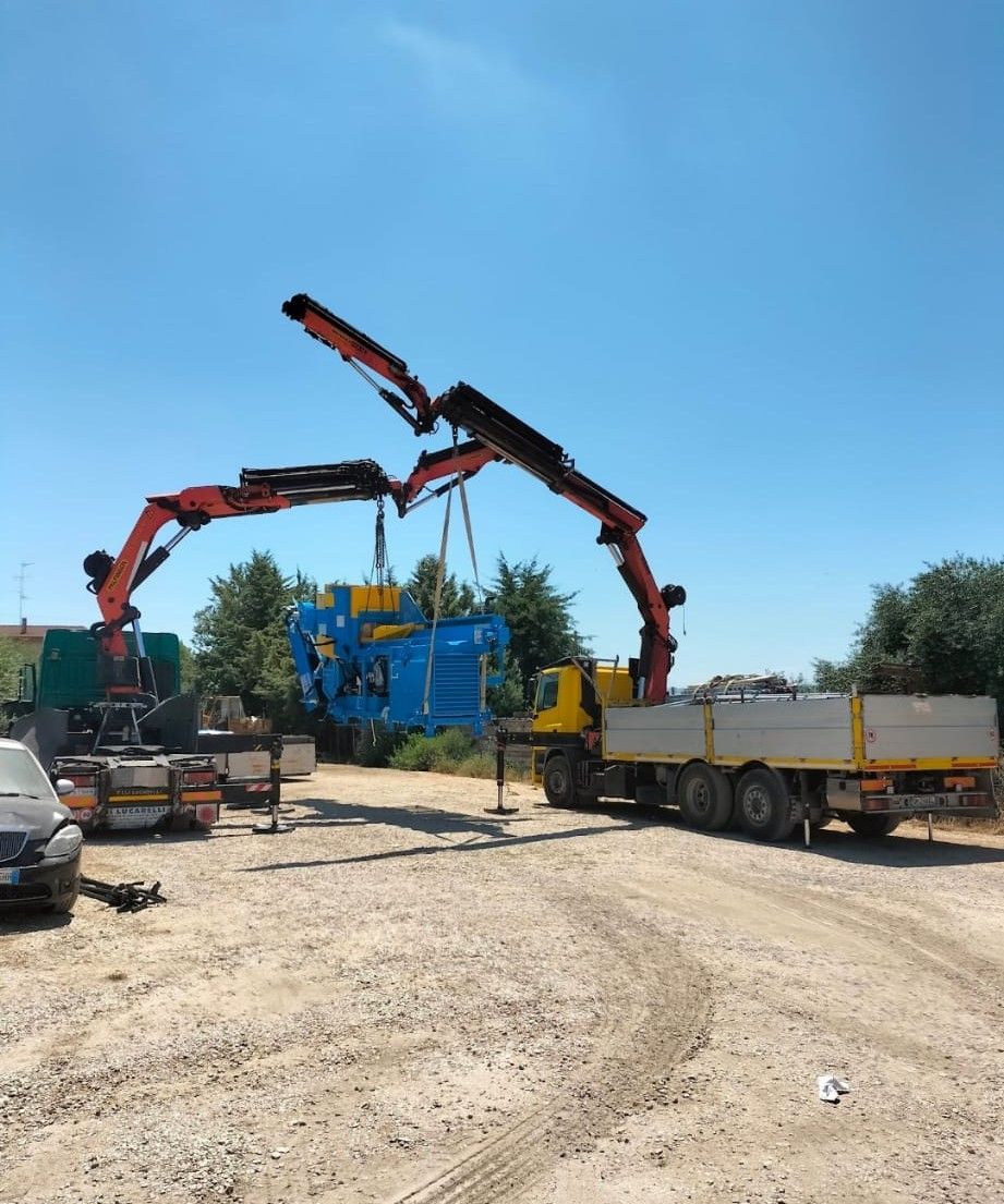 Cantiere edile con due autogru rosse che sollevano una struttura blu sotto un cielo sereno.