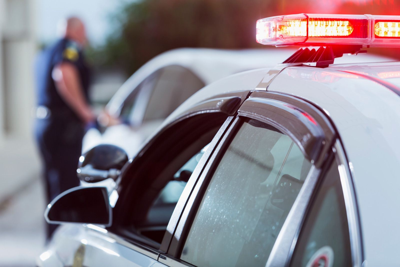 A police officer is standing next to a police car with a red light on top.