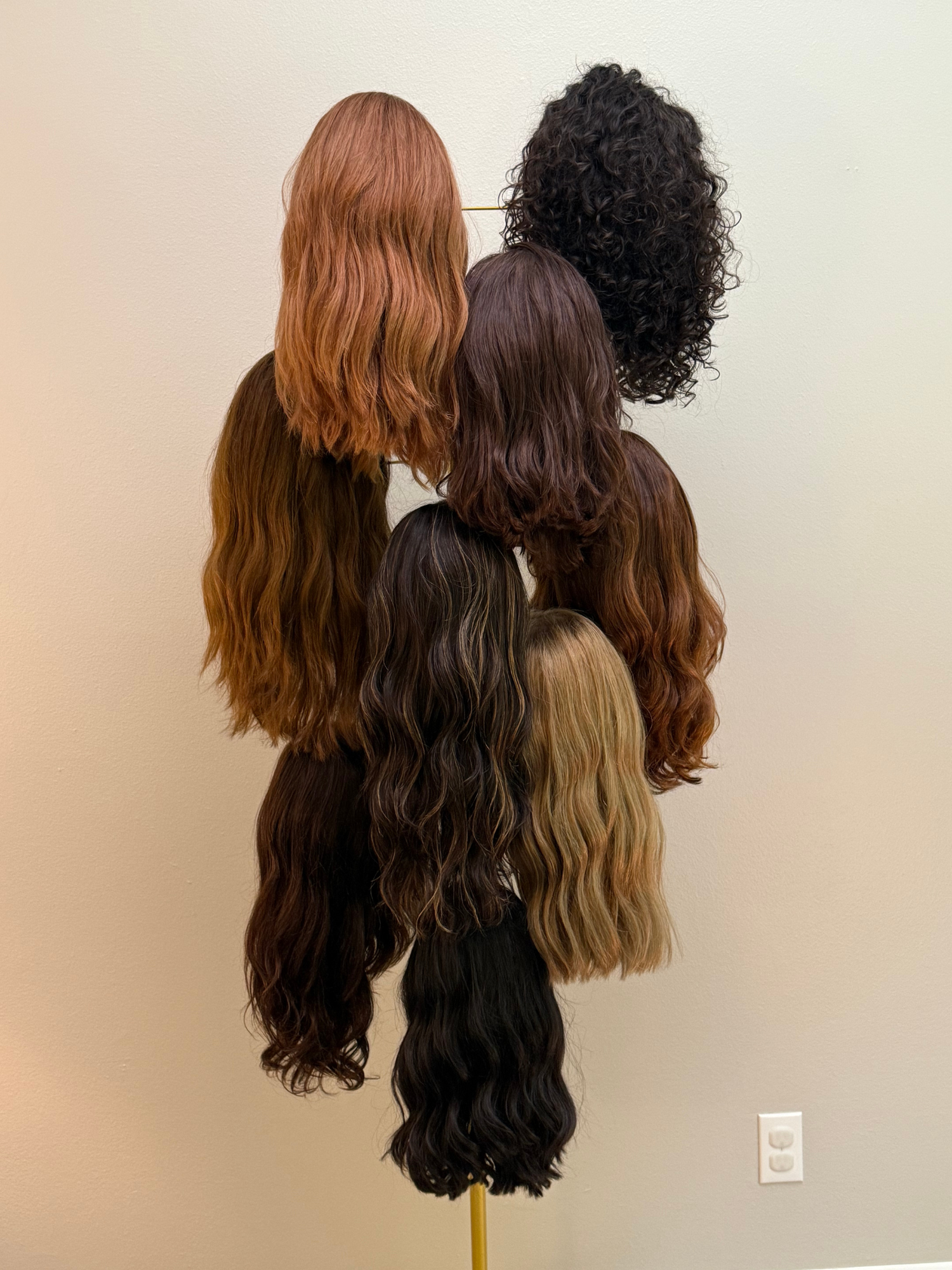 Wigs of various colors and styles hanging on a gold metal stand against a light-colored wall.