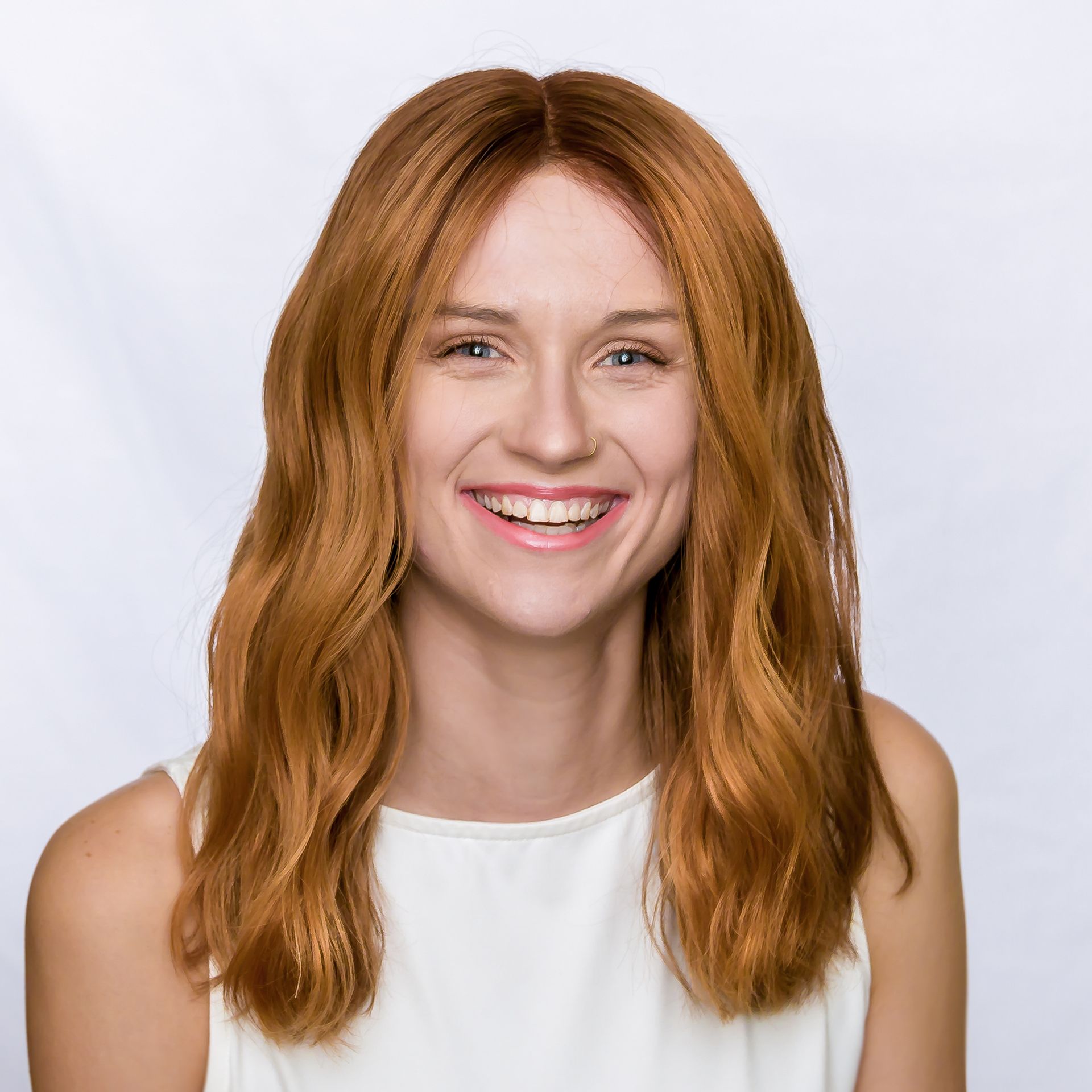 Woman with long brown hair and a friendly smile, wearing a white top, stands outside in front of green foliage.