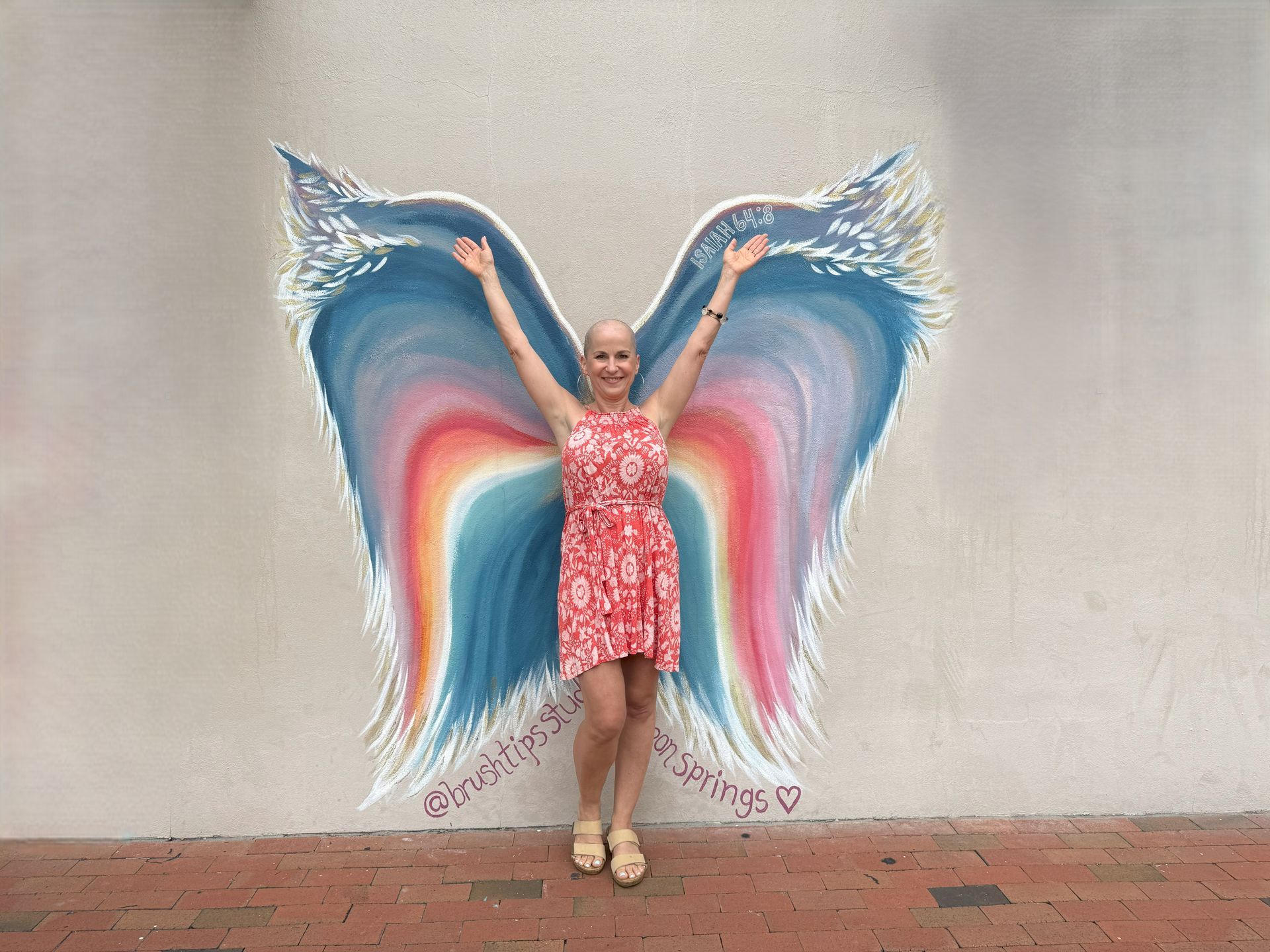 Woman stands in front of colorful angel wing mural, arms raised in joy.