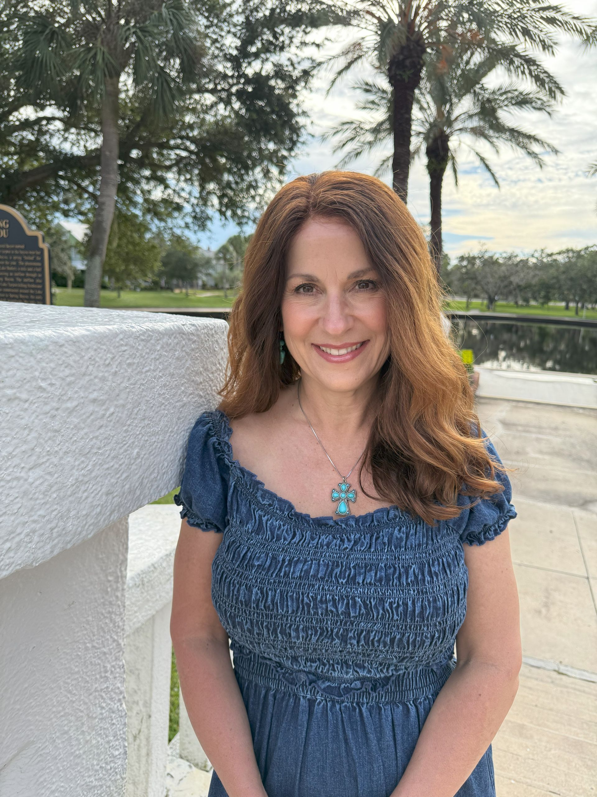 Woman with auburn hair smiles, wearing a blue dress, outdoors by a stone wall with palm trees and water in the background.