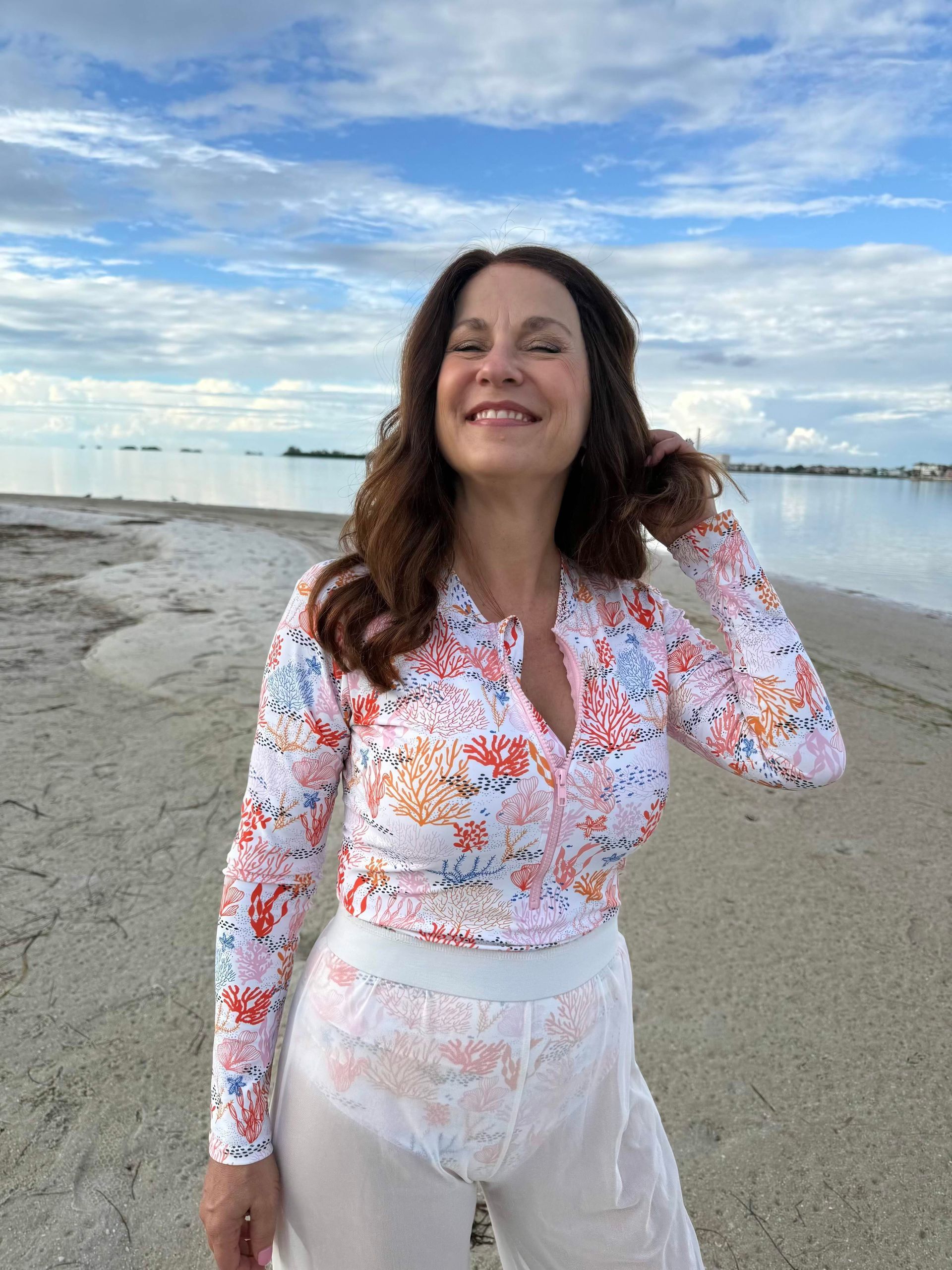 Woman smiling on a beach, wearing a floral top and white pants, with a calm sea and cloudy sky.