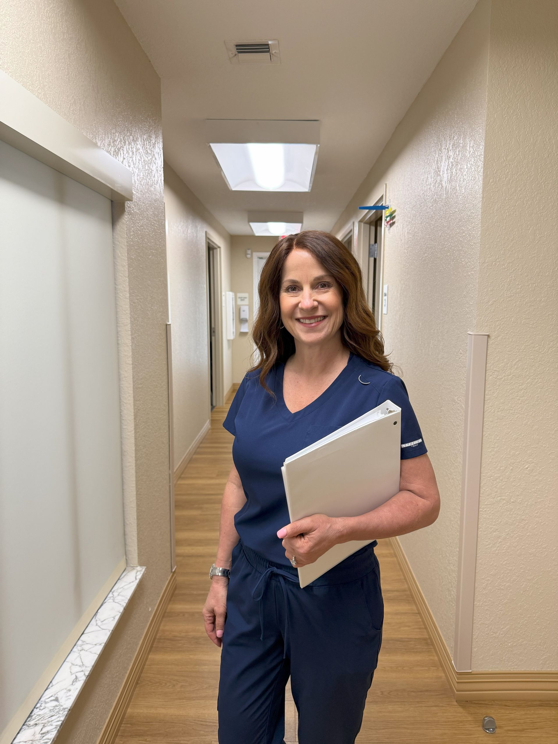 Woman in navy scrubs holding a file, standing in a hallway, smiling.