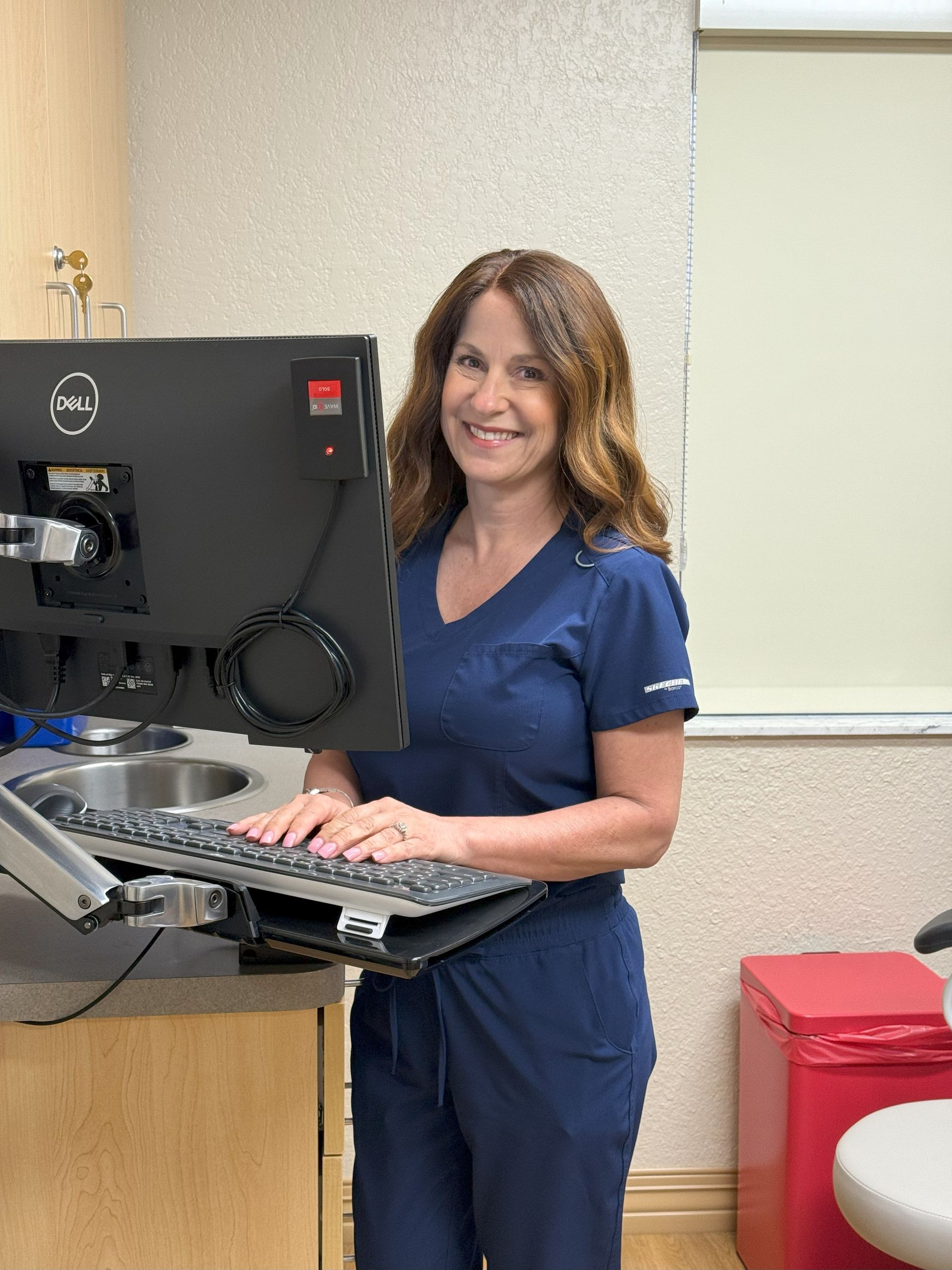 Woman in blue scrubs at a computer, smiling.  Medical office setting with sink and red disposal bin.