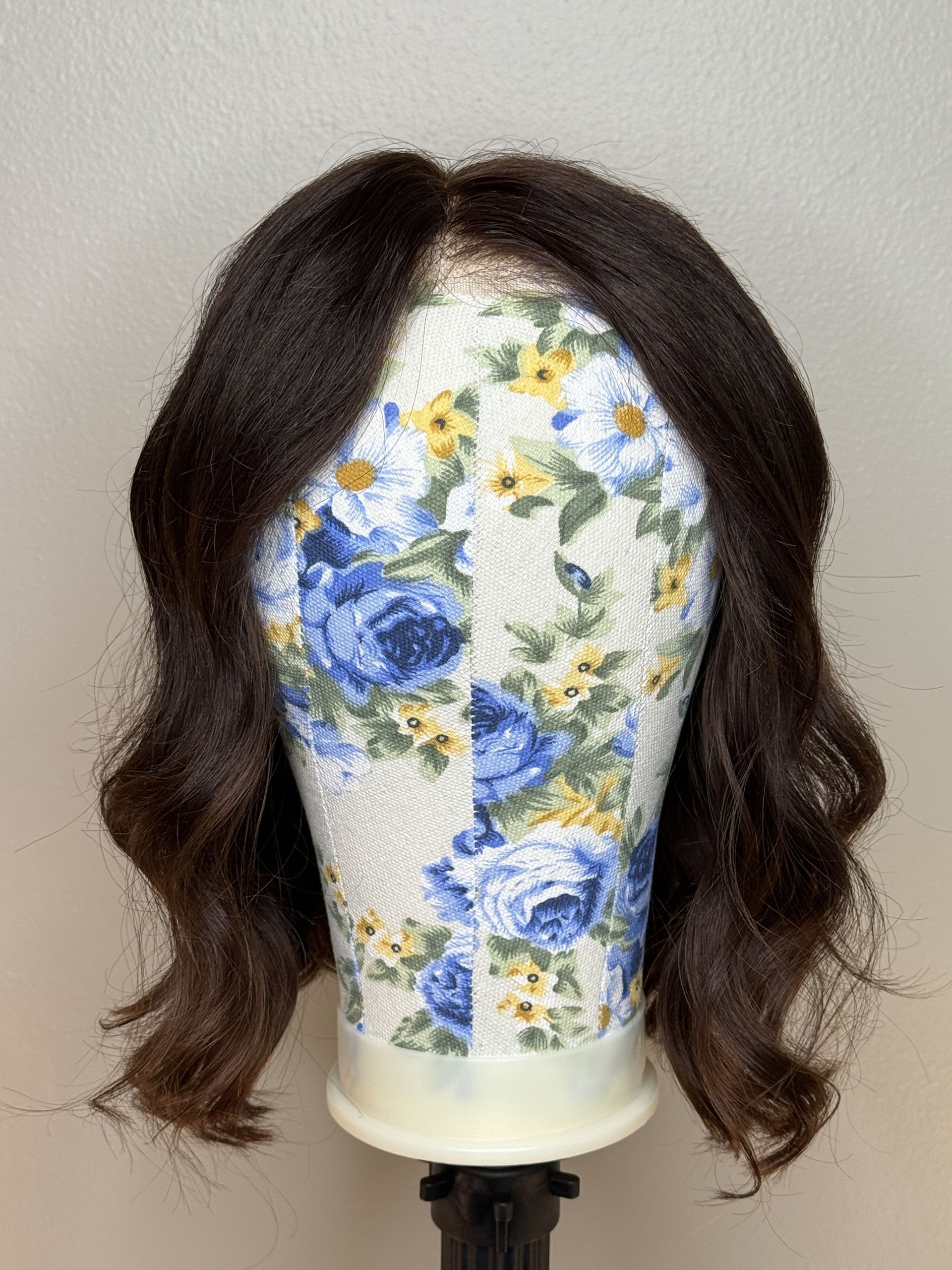 Brown curly wig styled on a flowered wig head, against a beige wall.