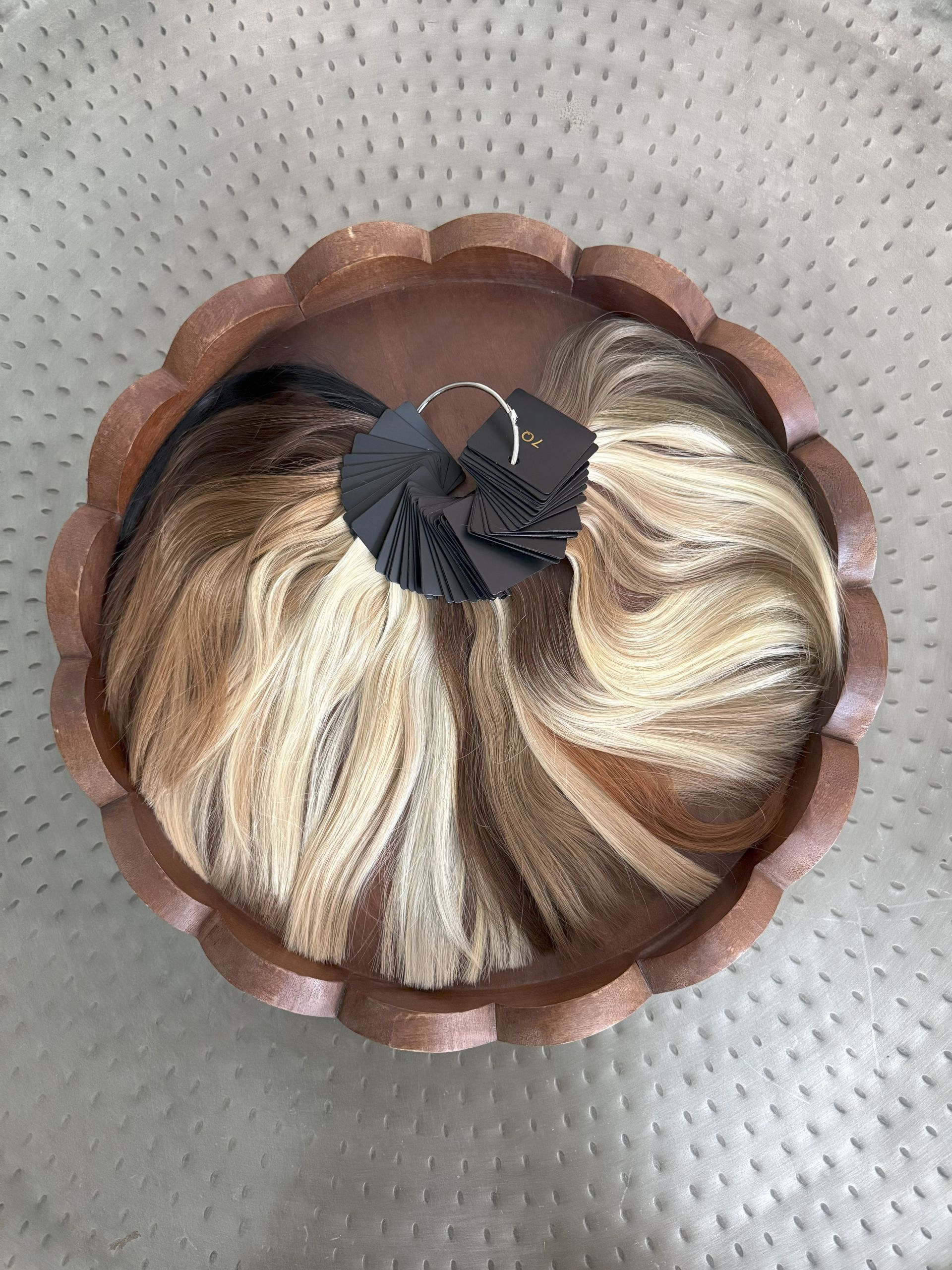 A wig with blonde and brown hair, in a decorative brown scalloped container, on a grey surface.