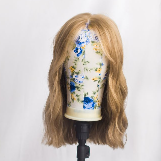 Woman with brown hair wearing a wig, smiling, against a white paneled backdrop. She is wearing a black top.