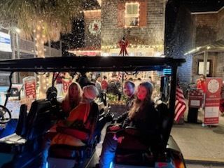 People ride in a golf cart decorated with lights, a snowy scene, buildings with Christmas decor.