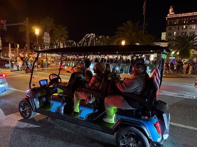 Golf cart with passengers driving on a street at night, with a crowd and buildings in the background.