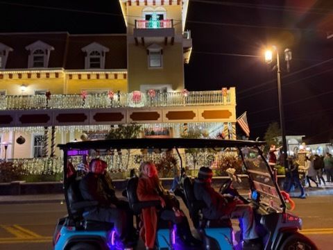 Golf cart with people, driving on a street with Christmas lights. Building in background.
