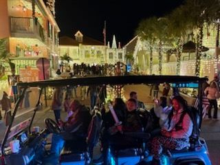 A golf cart with passengers on a nighttime holiday street, illuminated by lights and people.