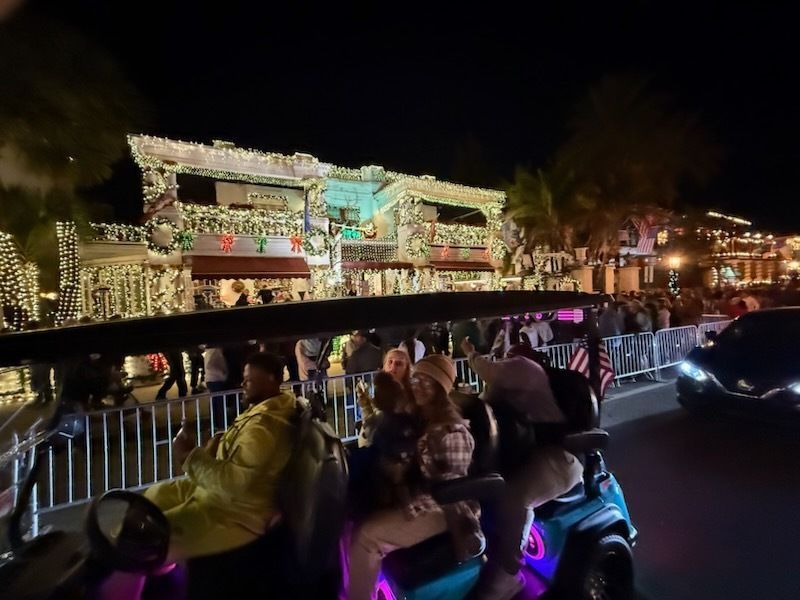 People ride in a golf cart at night, viewing buildings lit up with Christmas lights.
