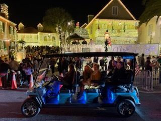A golf cart with passengers driving on a street decorated with lights at night.