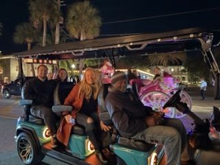 People on a teal golf cart, driver in front, others in seats. Palm trees in background, night.