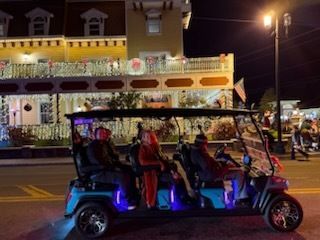 A golf cart with several people, illuminated in blue, driving at night in front of a building decorated with Christmas lights