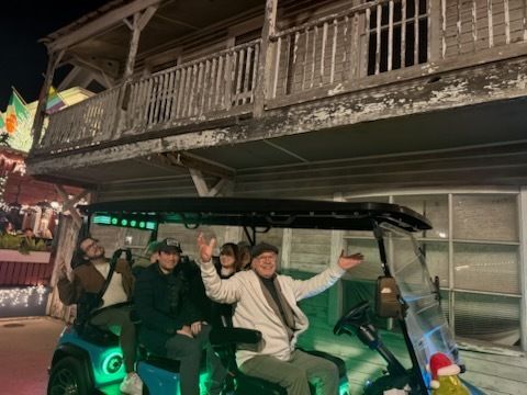 Group of people in a golf cart, smiling under a balcony at night, lit with festive lights.