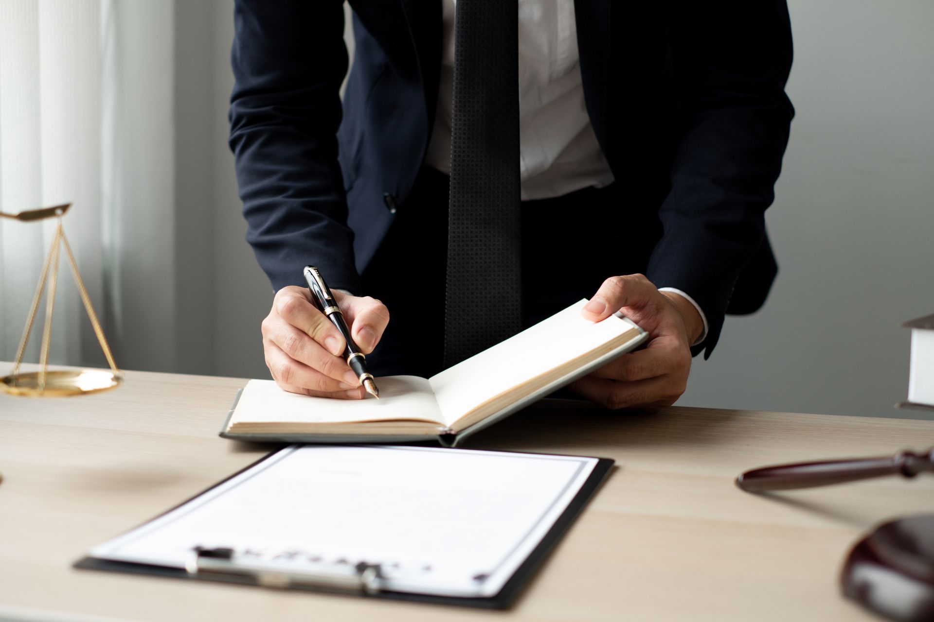 Lawyer in suit writing in a notebook at a desk with scales of justice and gavel nearby. Lawyer in suit writing in a notebook at a desk with scales of justice and gavel nearby.