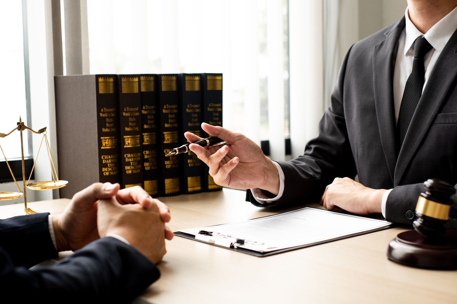 Lawyer in suit gestures while speaking to client at desk with documents and law books.