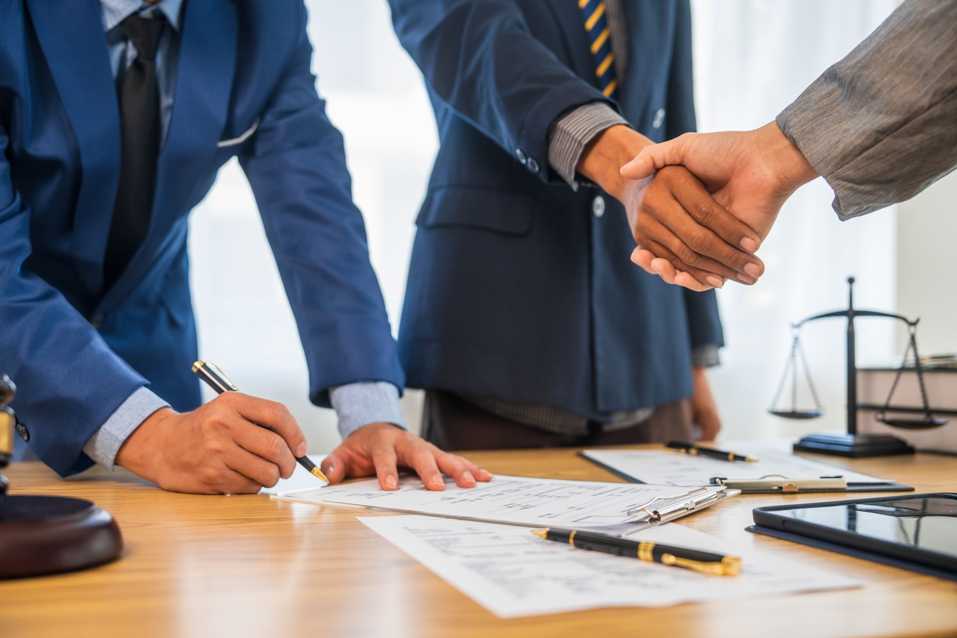 Three people in suits, one signing papers at a desk as two others shake hands.