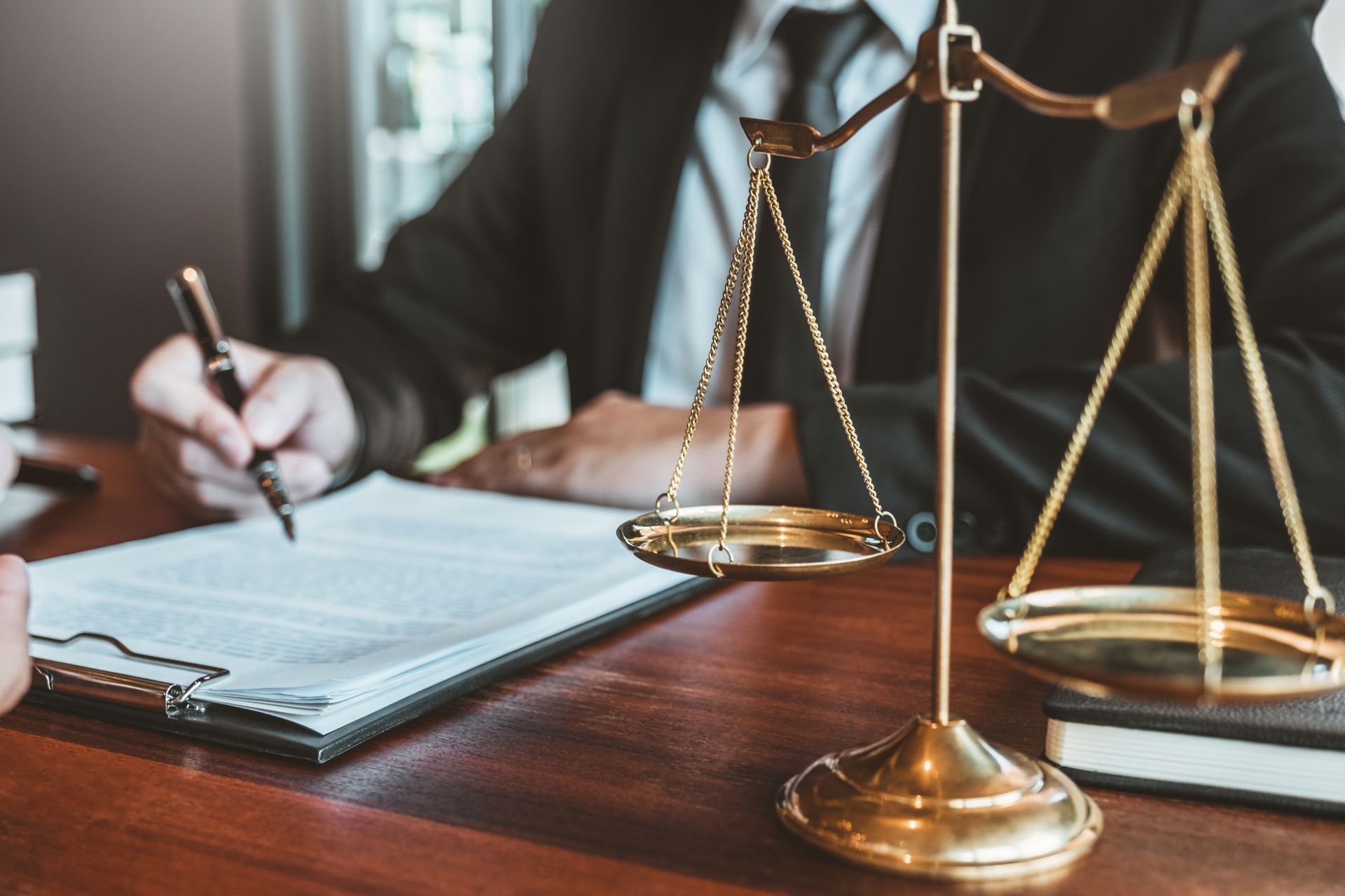 A legal professional in a suit writes on a document next to a brass balance scale of justice on a wooden desk.