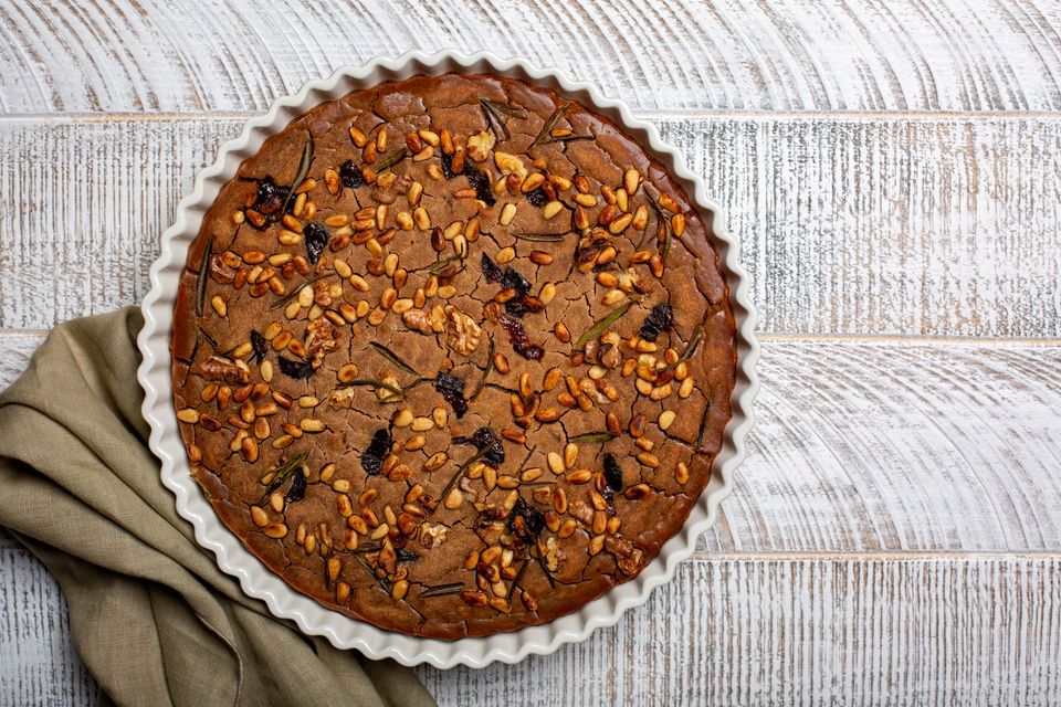 Torta rotonda con noci ed erbe aromatiche su un piatto con bordo arricciato, adagiata su una superficie di legno chiaro con un panno piegato.