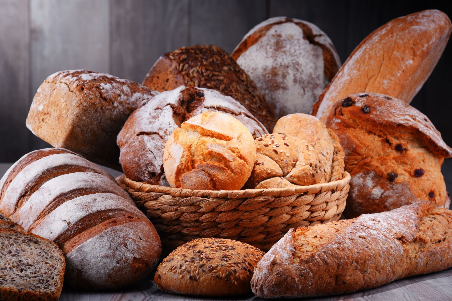 Assortimento di pane appena sfornato, tra cui pagnotte e panini, in un cestino e sparsi in giro.