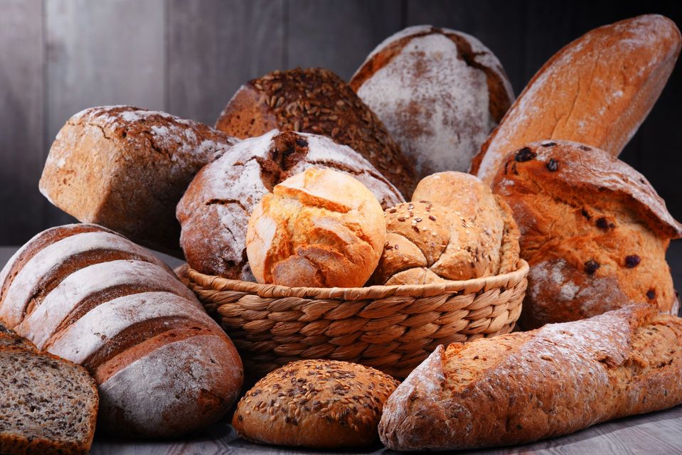 Assortimento di pane appena sfornato, tra cui pagnotte e panini, in un cestino e sparsi in giro.
