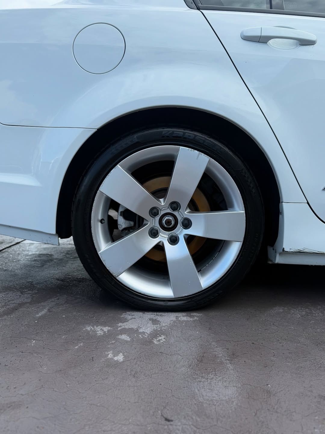 White car's rear wheel, flat tire on pavement, showing silver wheel, and faded brake disc.