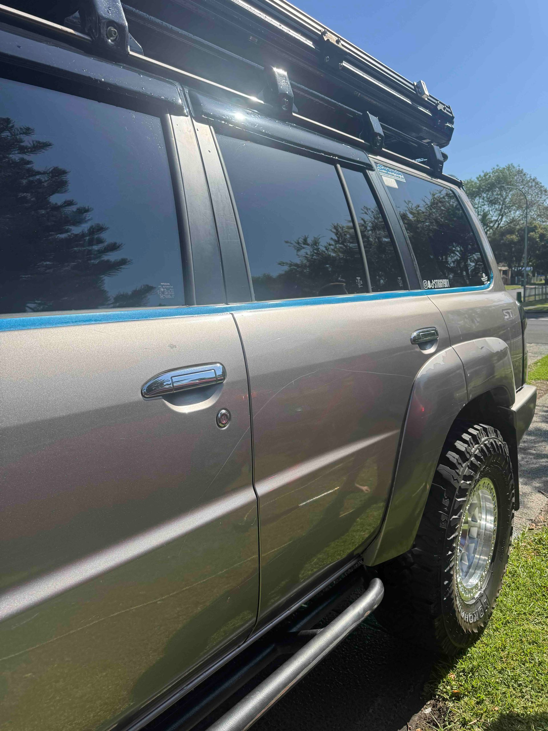 Gray SUV parked on grass, with dark tinted windows, a roof rack, and off-road tires, on a sunny day.