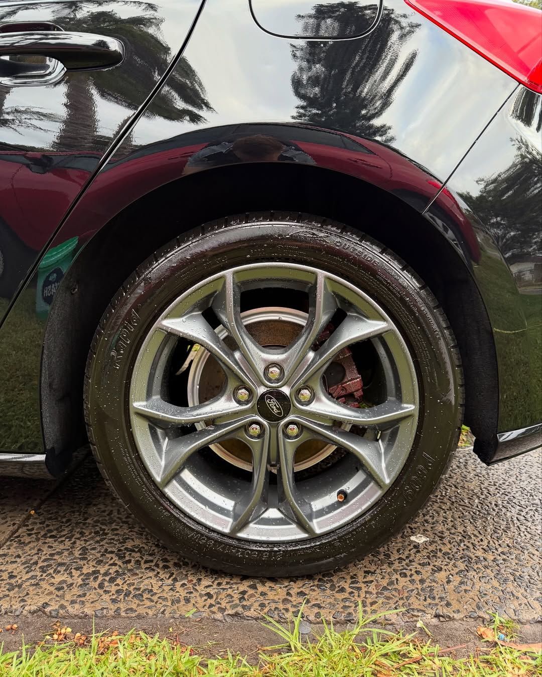 Black car's rear wheel with a gray rim and the Ford logo. Wet pavement, and a red taillight are visible.